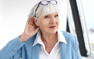 close up elegant stylish elderly woman in formal suit having hearing problems, holding hand at her ear, trying to hear you, saying: speak louder, please. age, maturity, people and health concept